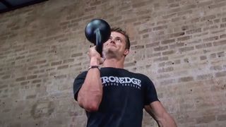 Man peforming a bottoms-up kettlebell press standing against a brick wall