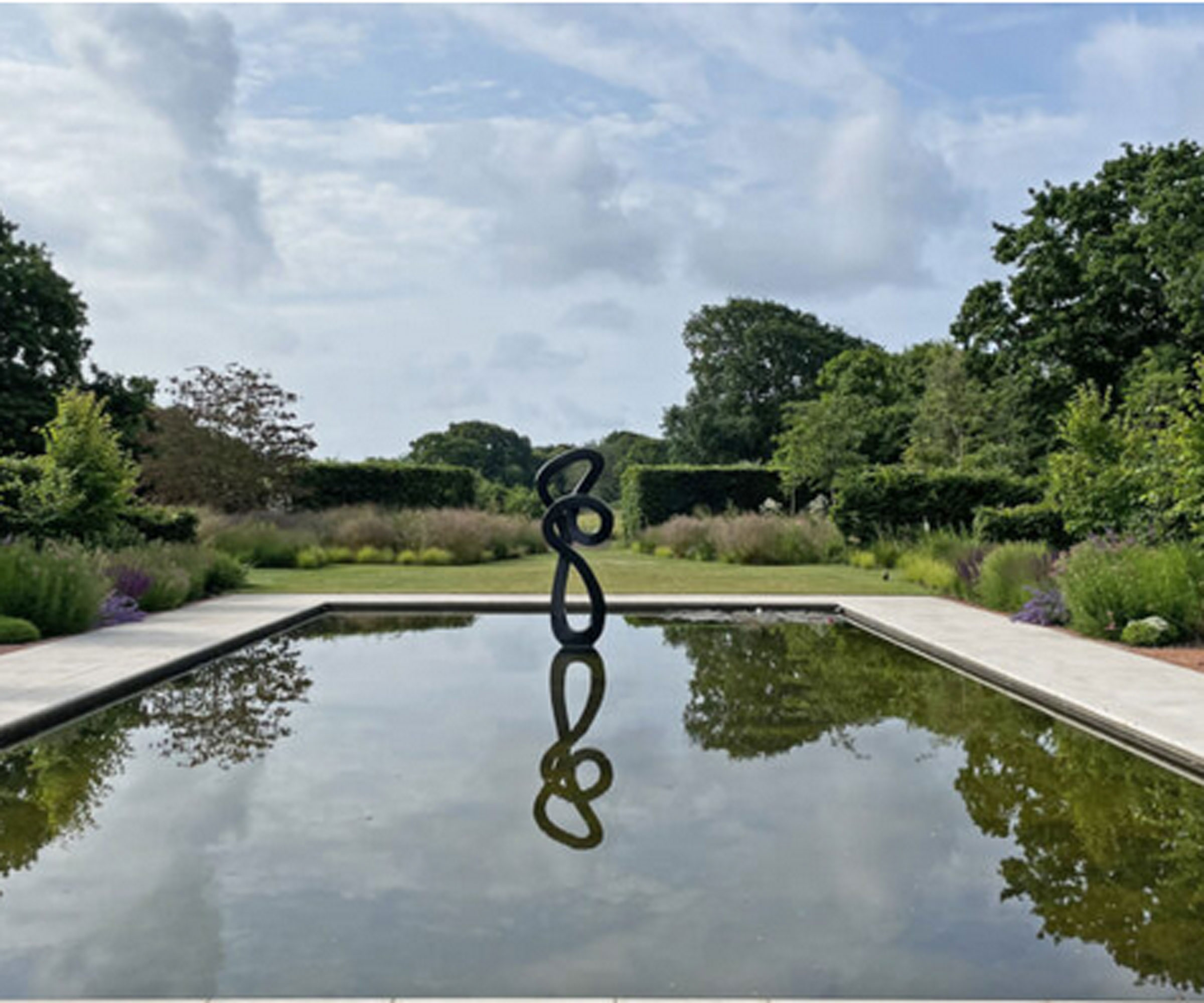 large modern garden pond with reflections and sculpture, surrounded by an edging of pavers, trees and lawn