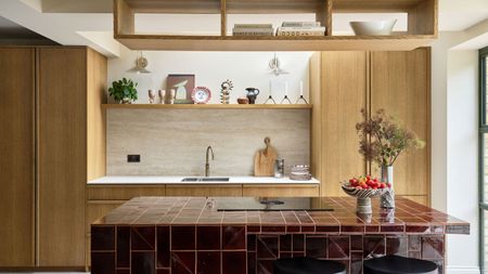 Kitchen with wooden cabinetry, a burgundy tiled island with a hob on the top, and a shelf of decor with glasses and artwork