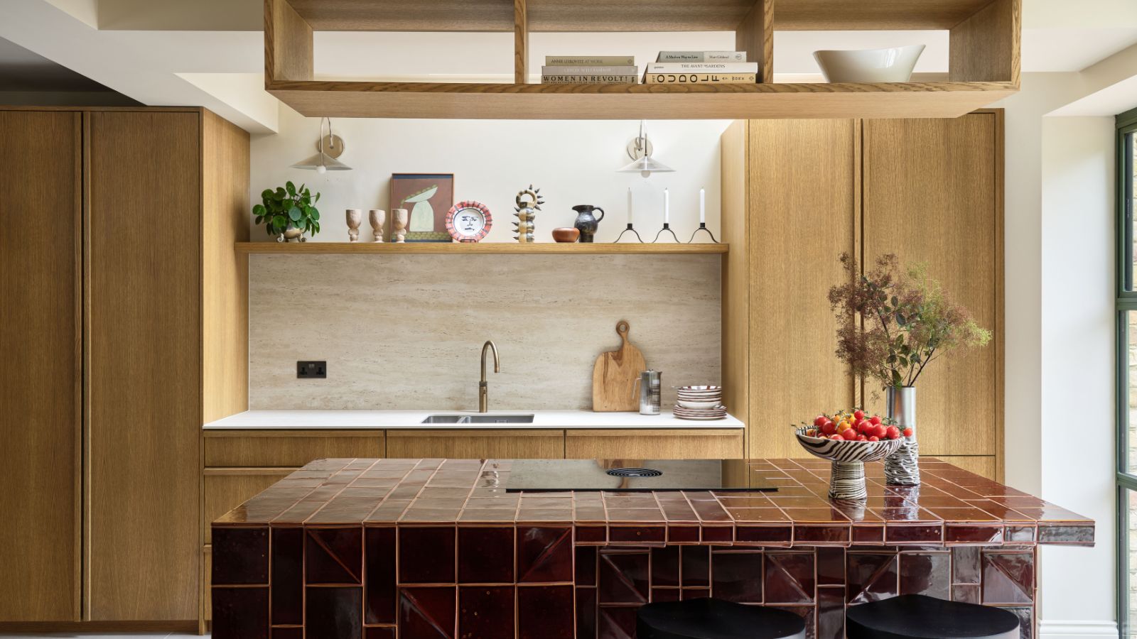 Kitchen with wooden cabinetry, a burgundy tiled island with a hob on the top, and a shelf of decor with glasses and artwork