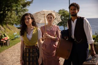 Grace Ambrose, Imogen Waterhouse and Matthew Broome walking with luggage and a parasol by the ocean in buccaneers season 2