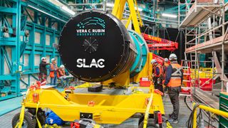 a massive black cylinder is lifted by crane into a round metal enclosure in a large warehouse