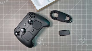 A sleek black Steam Controller with buttons and joysticks lies on a grid-patterned surface beside a coiled cable and a small puck charging accessory.