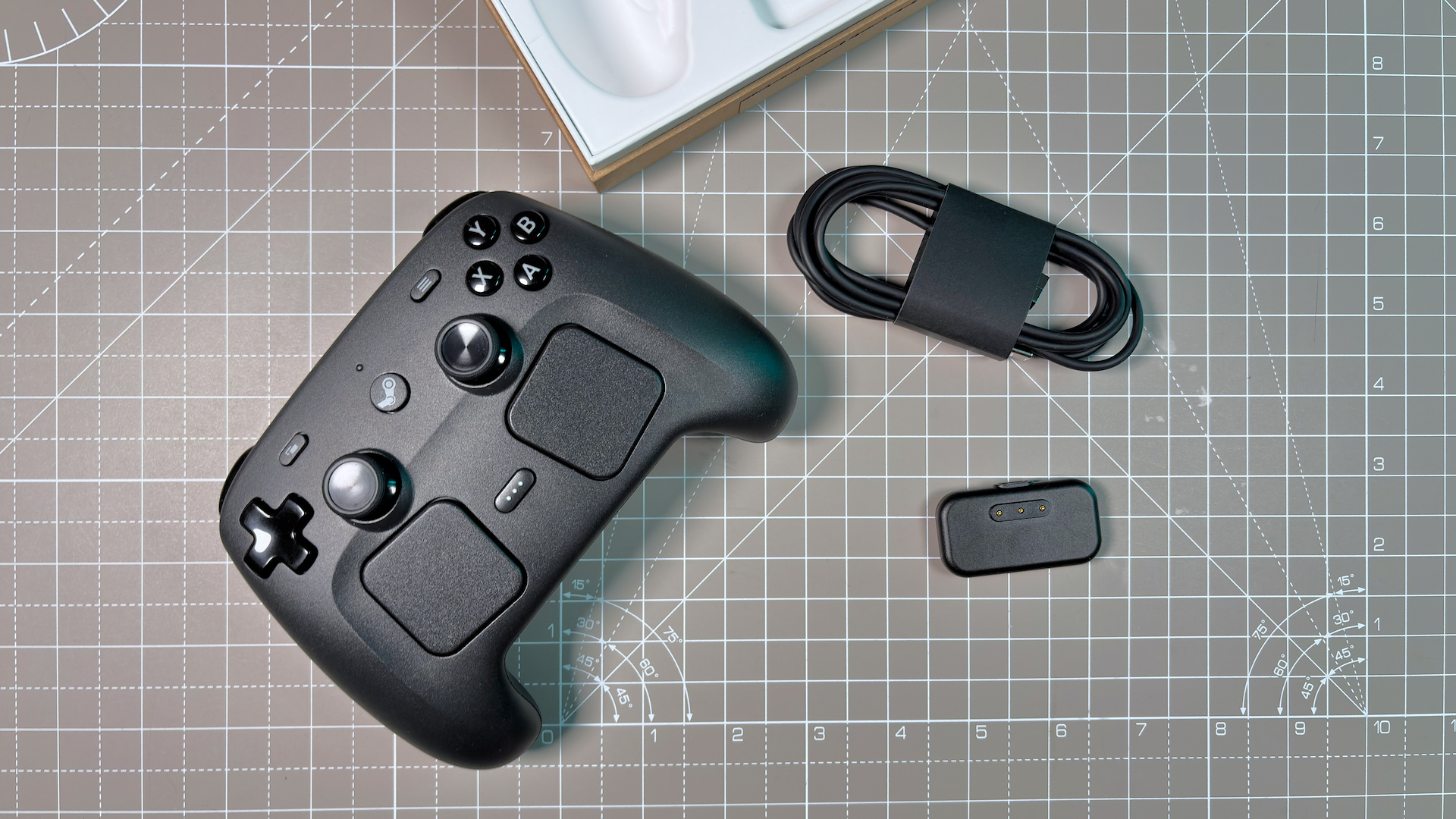 A sleek black Steam Controller with buttons and joysticks lies on a grid-patterned surface beside a coiled cable and a small puck charging accessory.