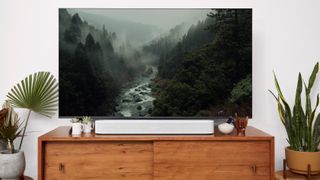 A white Sonos Beam Gen 2 on a wooden TV stand in front of a TV. On screen is a misty, wooded valley with a river running through it.