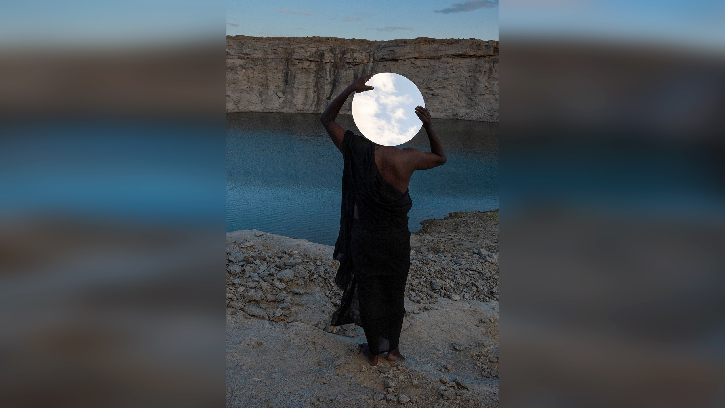 A person in a black dress stands at a rocky cliff's edge, holding a round mirror overhead that reflects a partly cloudy sky, facing a calm, blue lake