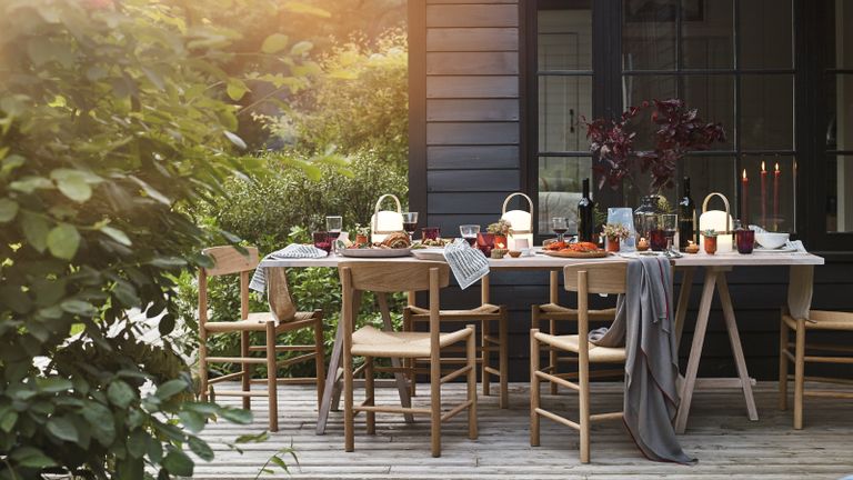 Outdoor dining ideas with wooden table and chairs outside black wood panelled house, with autumnal table decor and grey blankets