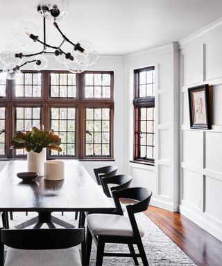 White dining room with paneled walls, hardwood flooring, rug under black table and chairs, globe glass pendant light above table, dark wooden window frames