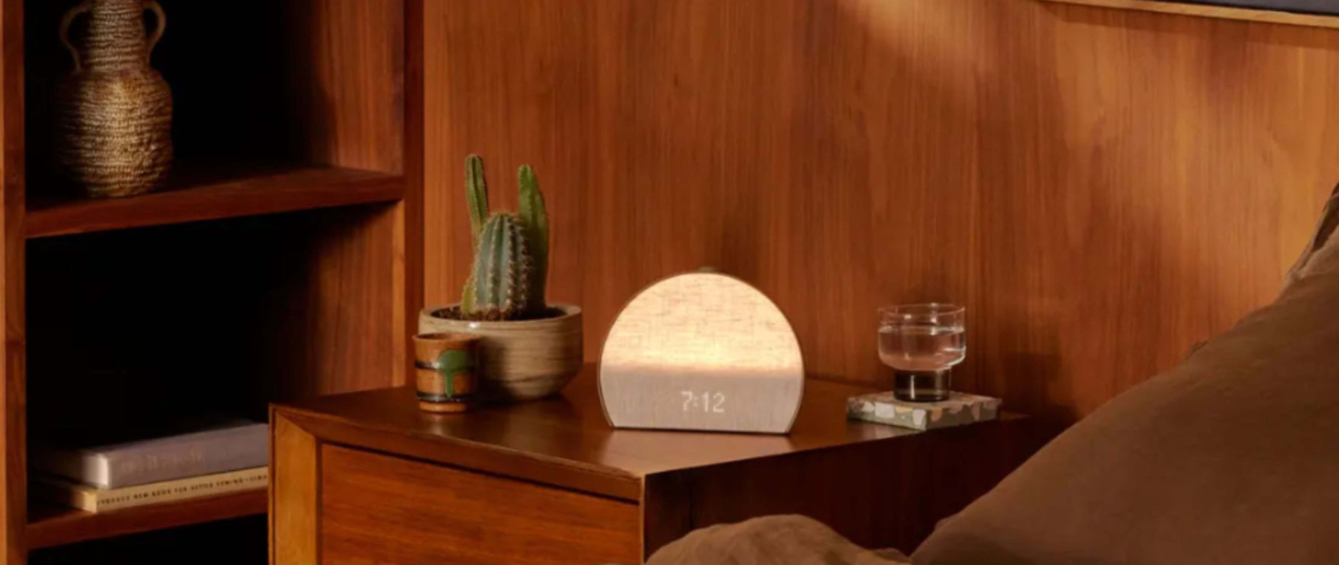 Hatch Restore 3 sunrise alarm clock on wooden bedside table next to cactus and glass of water with wooden wall behind