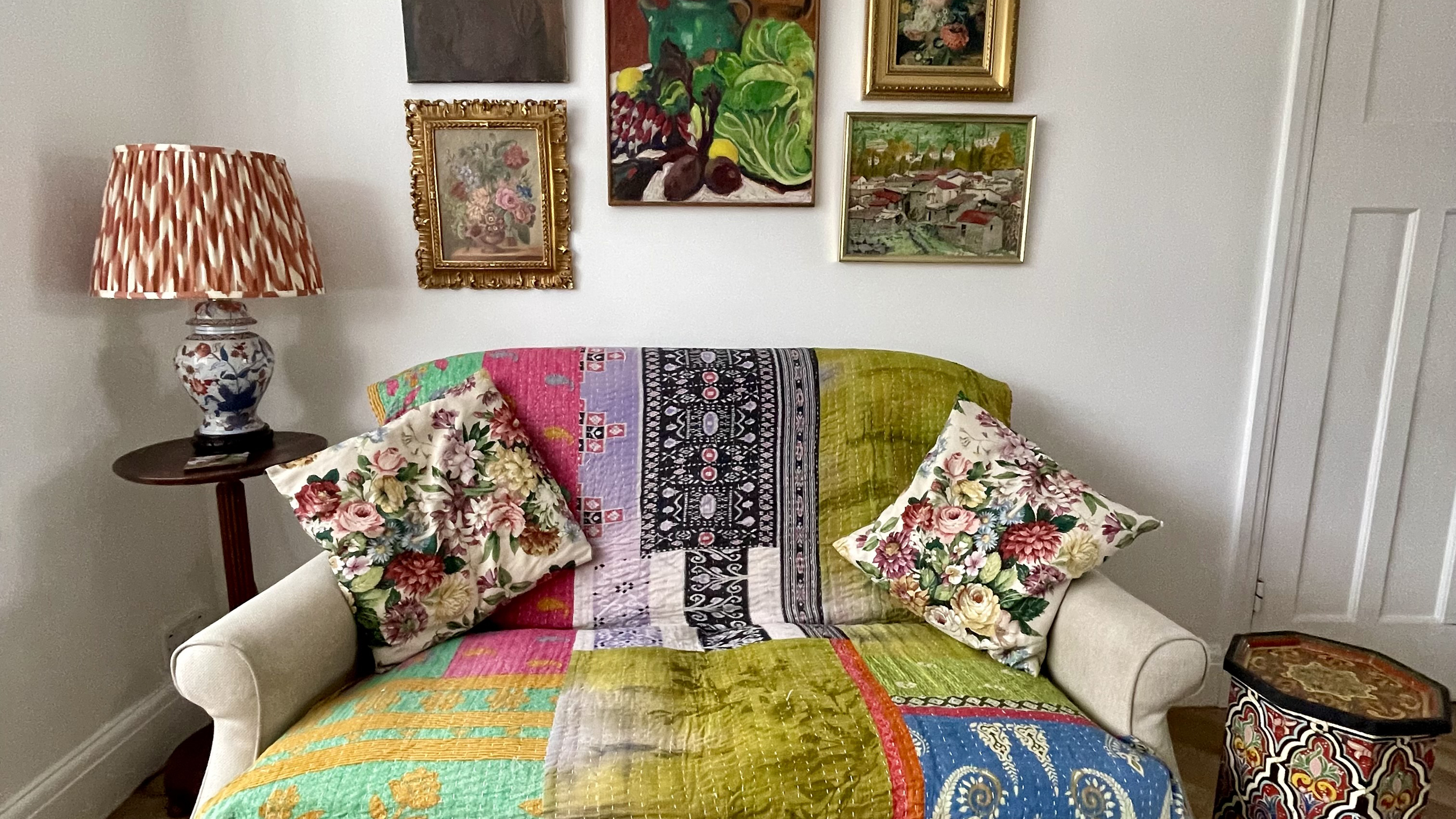 White living room with a small sofa covered with a colourful throw and vintage floral cushions, and a small gallery of painted artwork on the wall behind