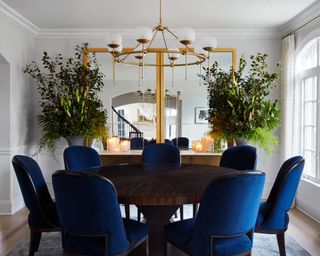 Royal blue padded dining chairs around dark walnut round dining table, with brass frame oversized wall mirrors and brass circular focal pendant.