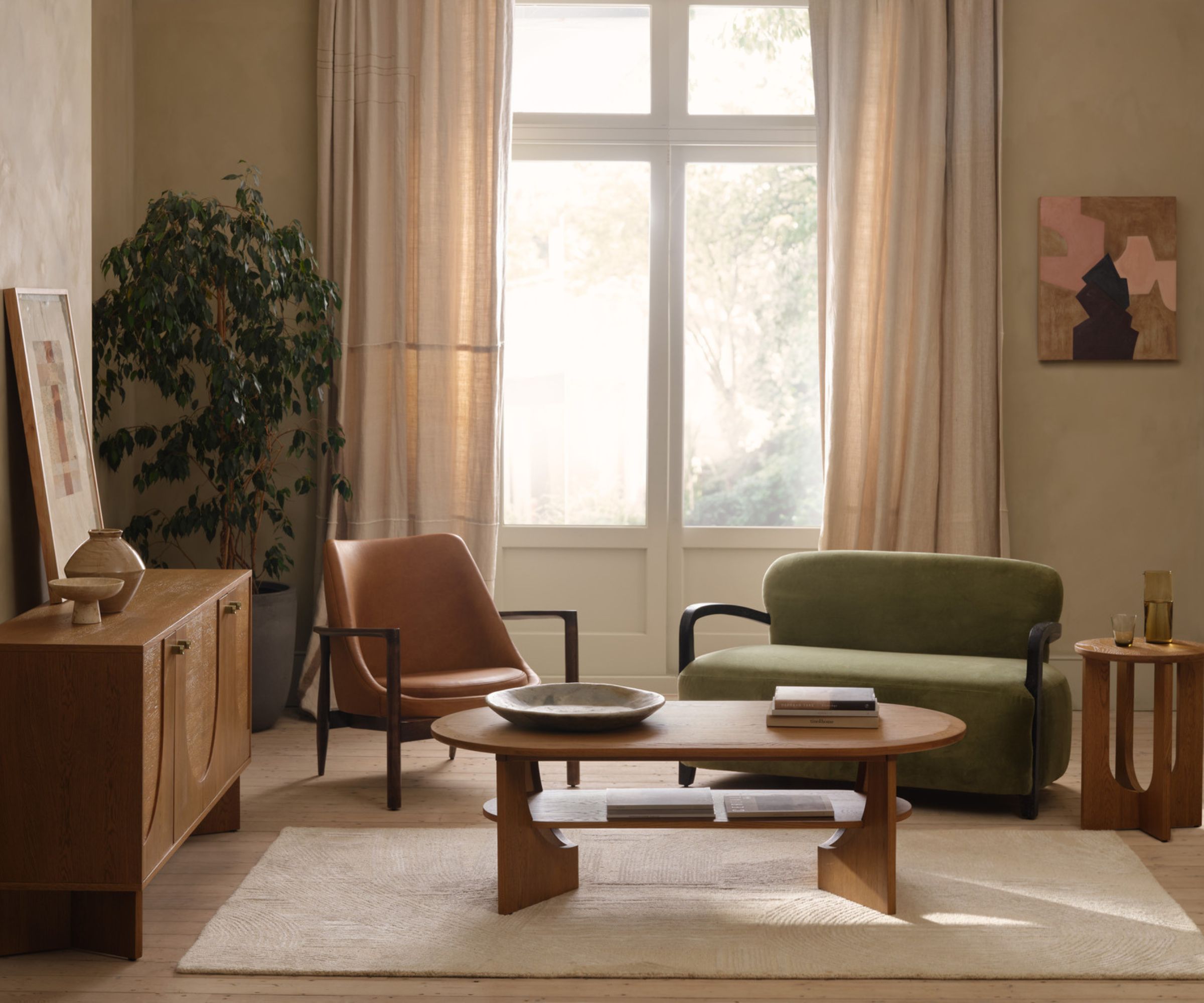 Green sofa, tan leather chair, oval coffee table and wooden sideboard in sunlit, earthy living room.