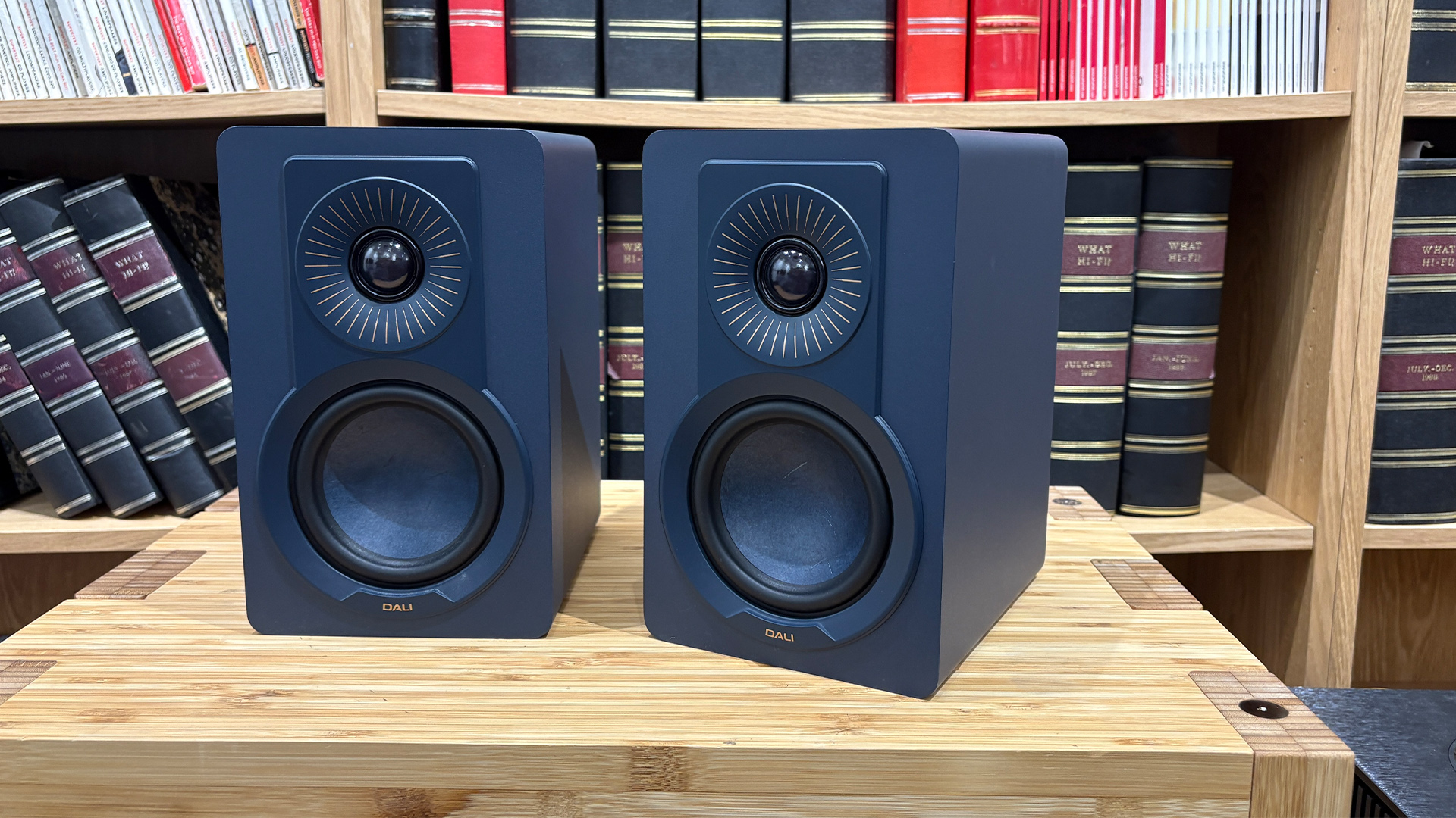 Dali Kupid standmount speakers on wooden hi-fi rack in front of bookcase
