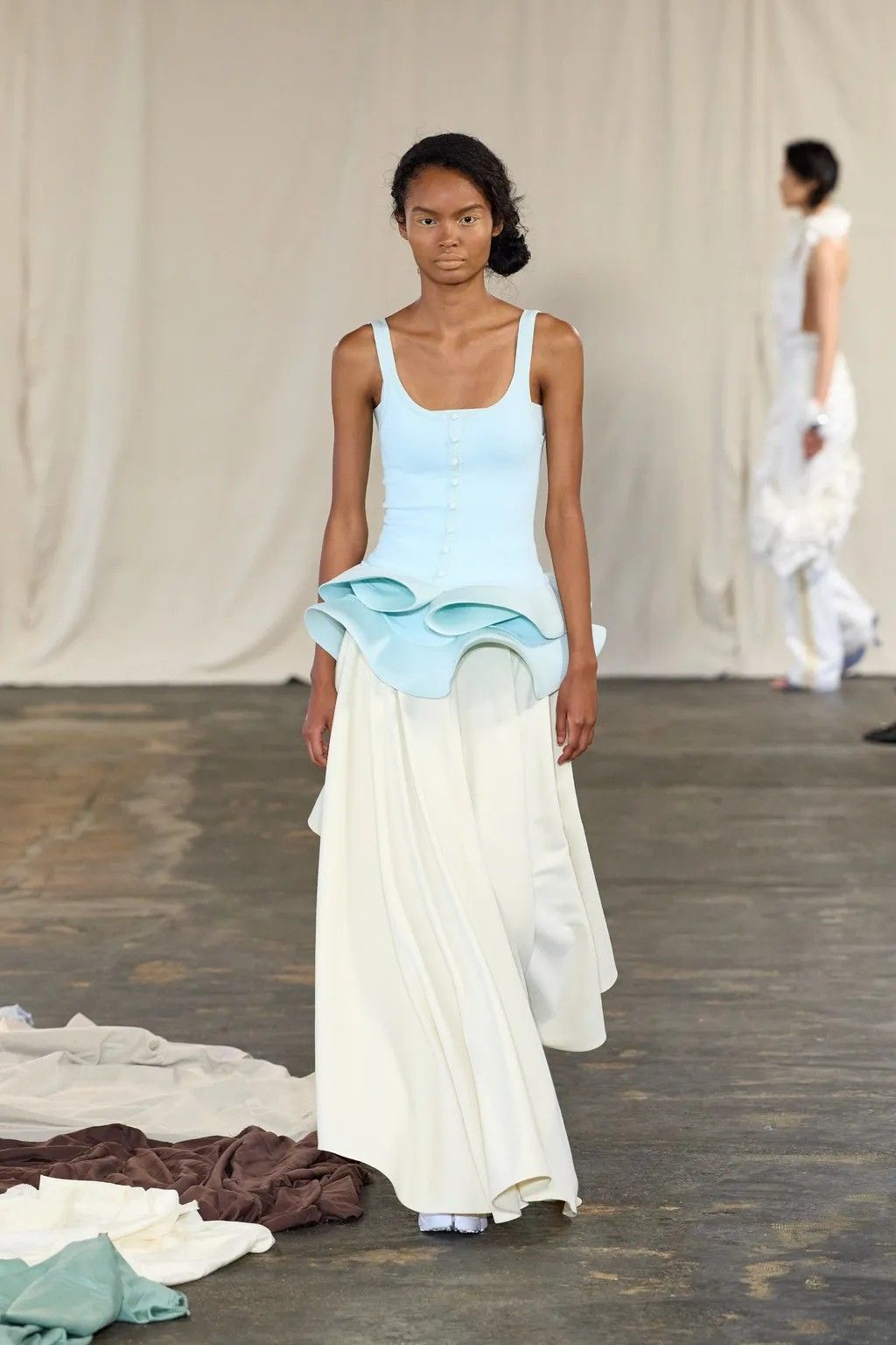 A woman wears a pale blue, sculptural top over a cream, loose-fitting skirt while walking down the runway with her hair gathered in a low bun.