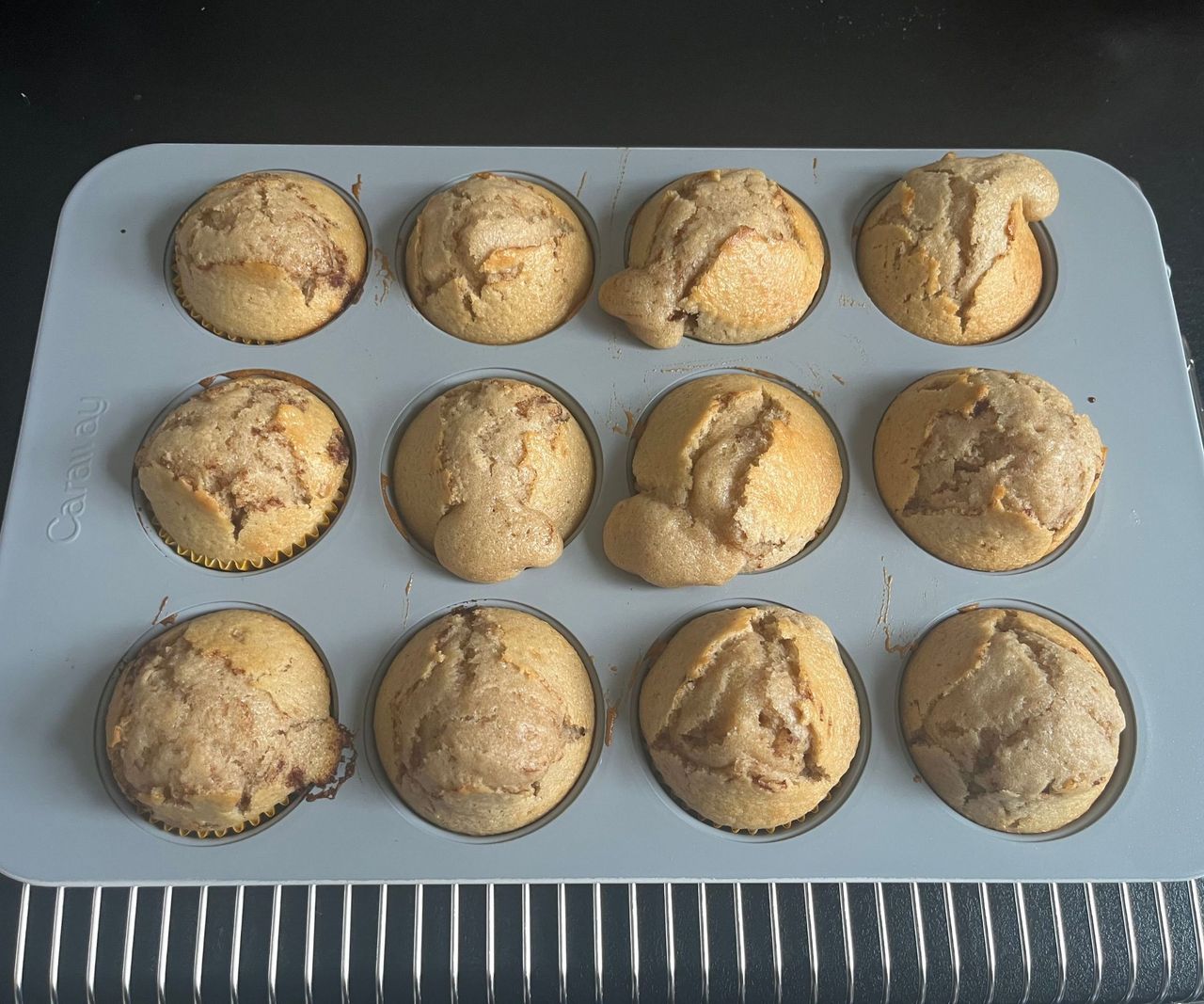 A grey muffin tin with 12 golden muffins inside on a wire cooling rack