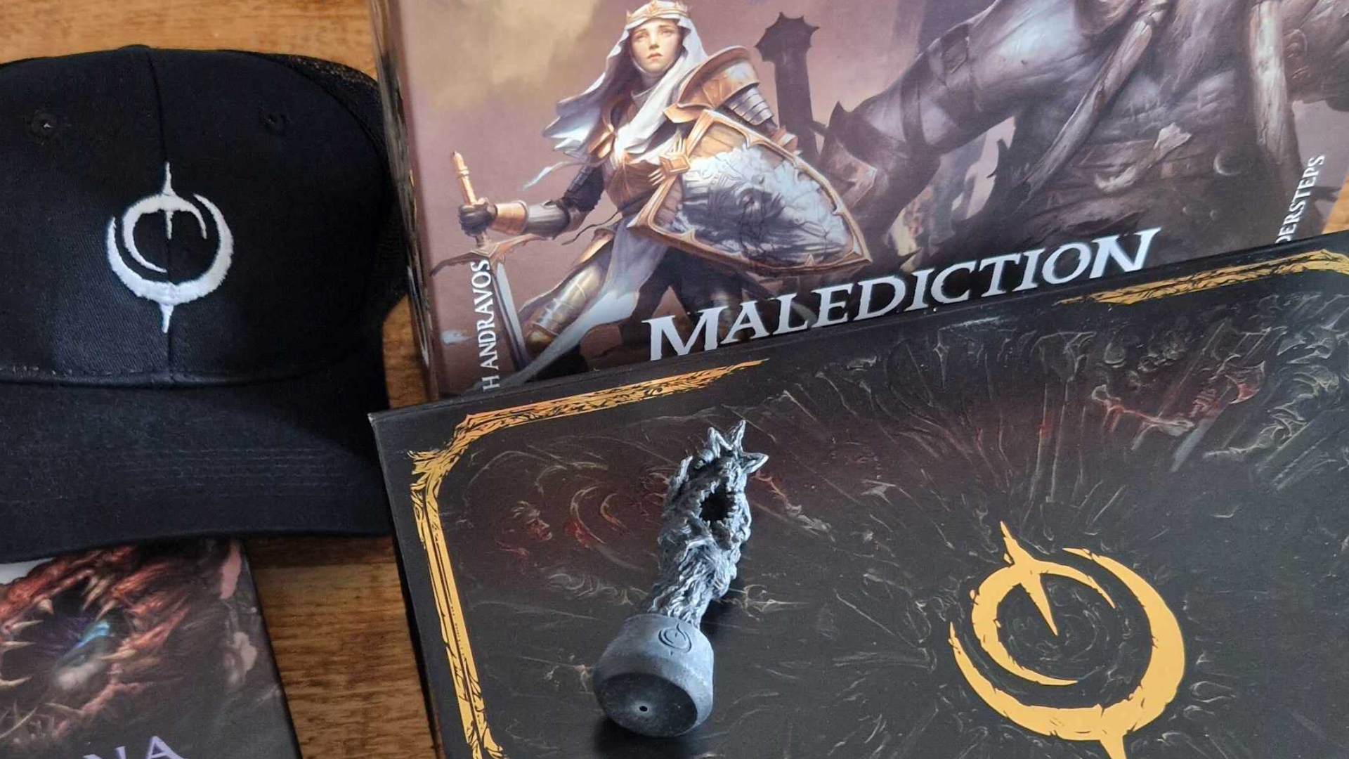 A Malediction two-player box set alongside a gold and black press pack, statue-esque figure, hat, and more on a wooden table