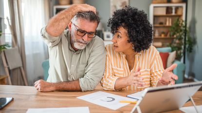 An older couple look shocked as they work on paperwork together at their dining room table.