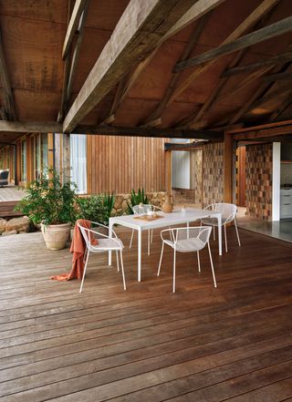 porch with white table and chairs, fall colored russet blanket, ceramic and wood accessories on the table, pots with plants, wooden floor, beams, view into kitchen