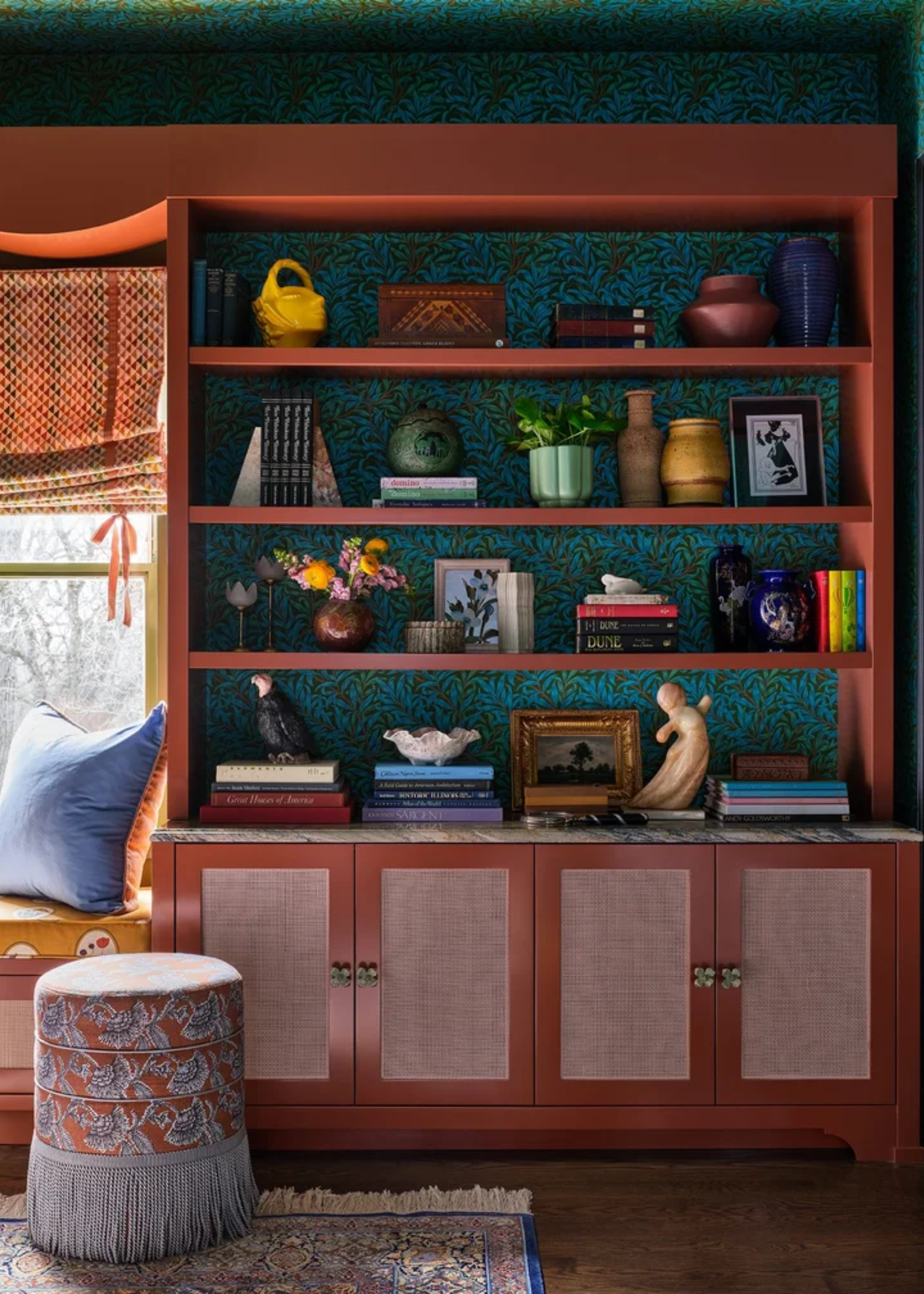A living room with green patterned wallpaper, an orange open display shelf with cabinets below holding vases, trinkets, frames, books and bowls. A fringed stool sits on a rug by a bay window.