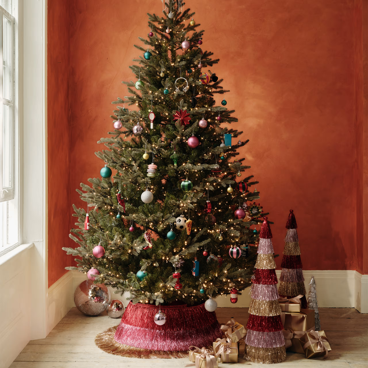 M&amp;S tinsel tree skirt in an living room with orange painted walls and wooden floorboards. There are two disco balls, two tinsel trees and a collection of gold presents at the base.