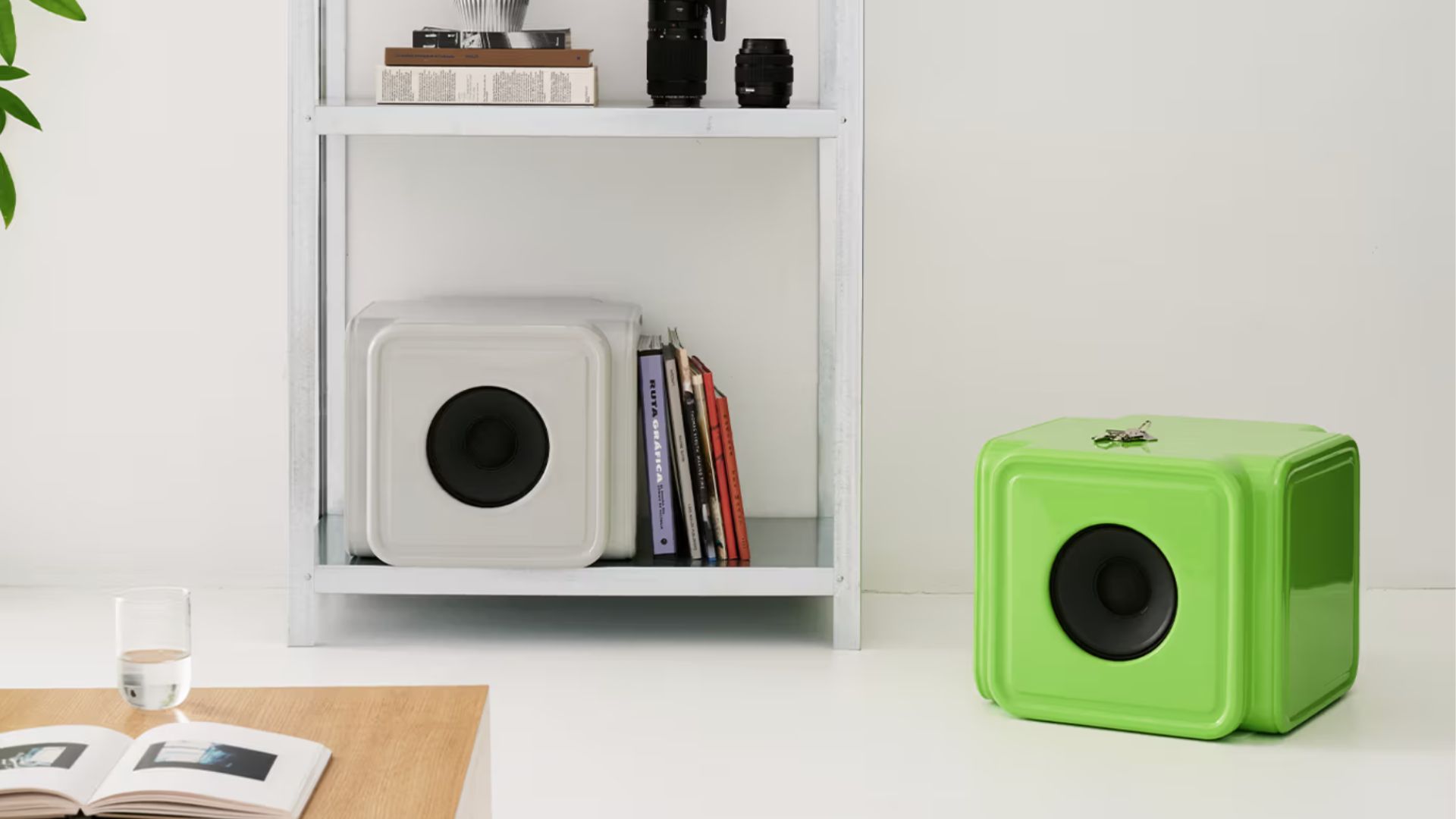 an image of the grey and green Caliper Alaska Alaska speakers. The grey one is on the bottom rung of white ladder-style shelving unit. The green one is adjacent to is on the floor