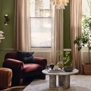 A living room with olive green-painted walls, semi-sheer beige curtains framing the windows and a burgundy accemt chair positioned in front of it