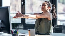 A woman in a grey top and glasses stretches her arms in front of her while sat at a desk