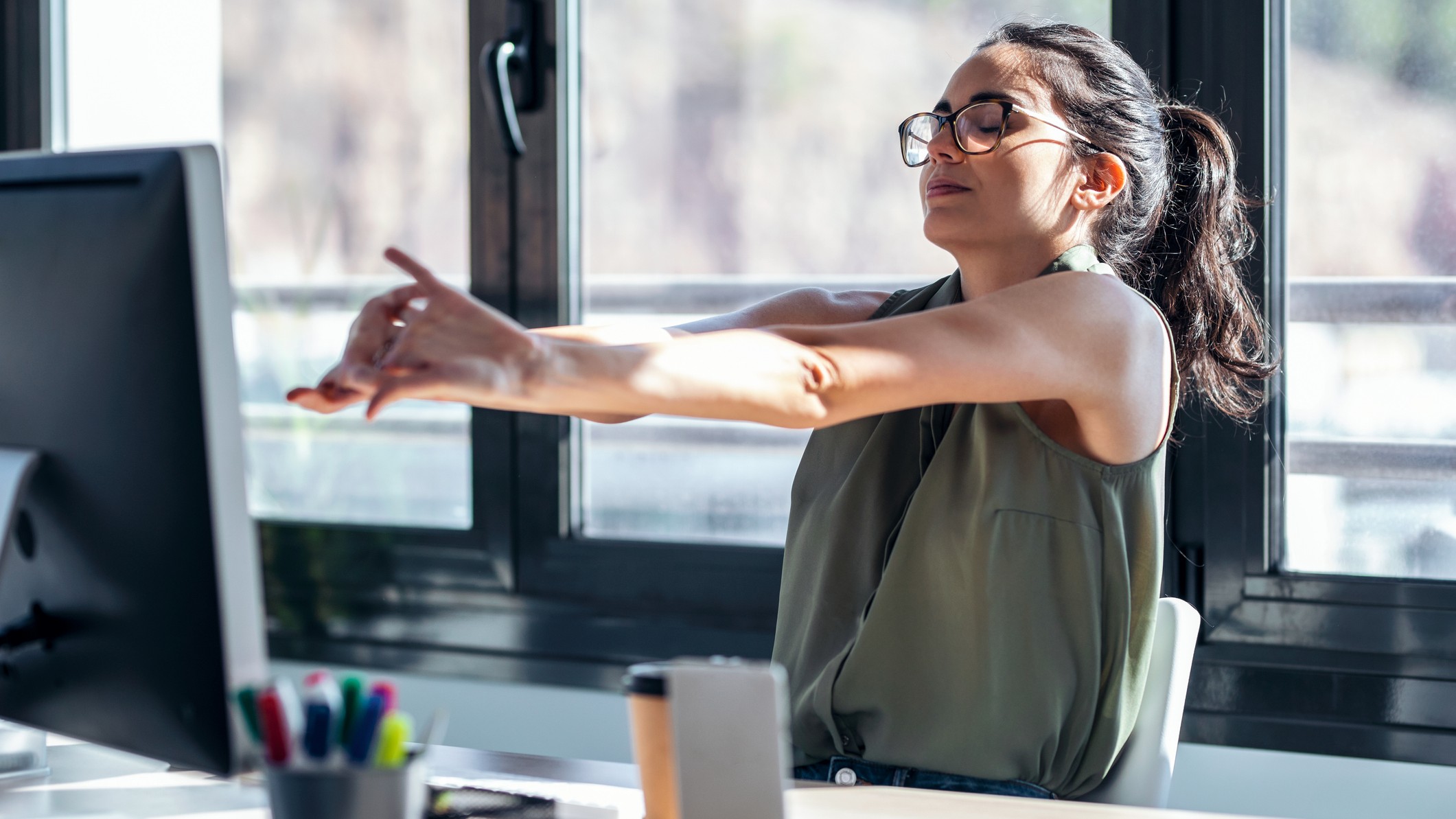 A woman in a grey top and glasses stretches her arms in front of her while sat at a desk