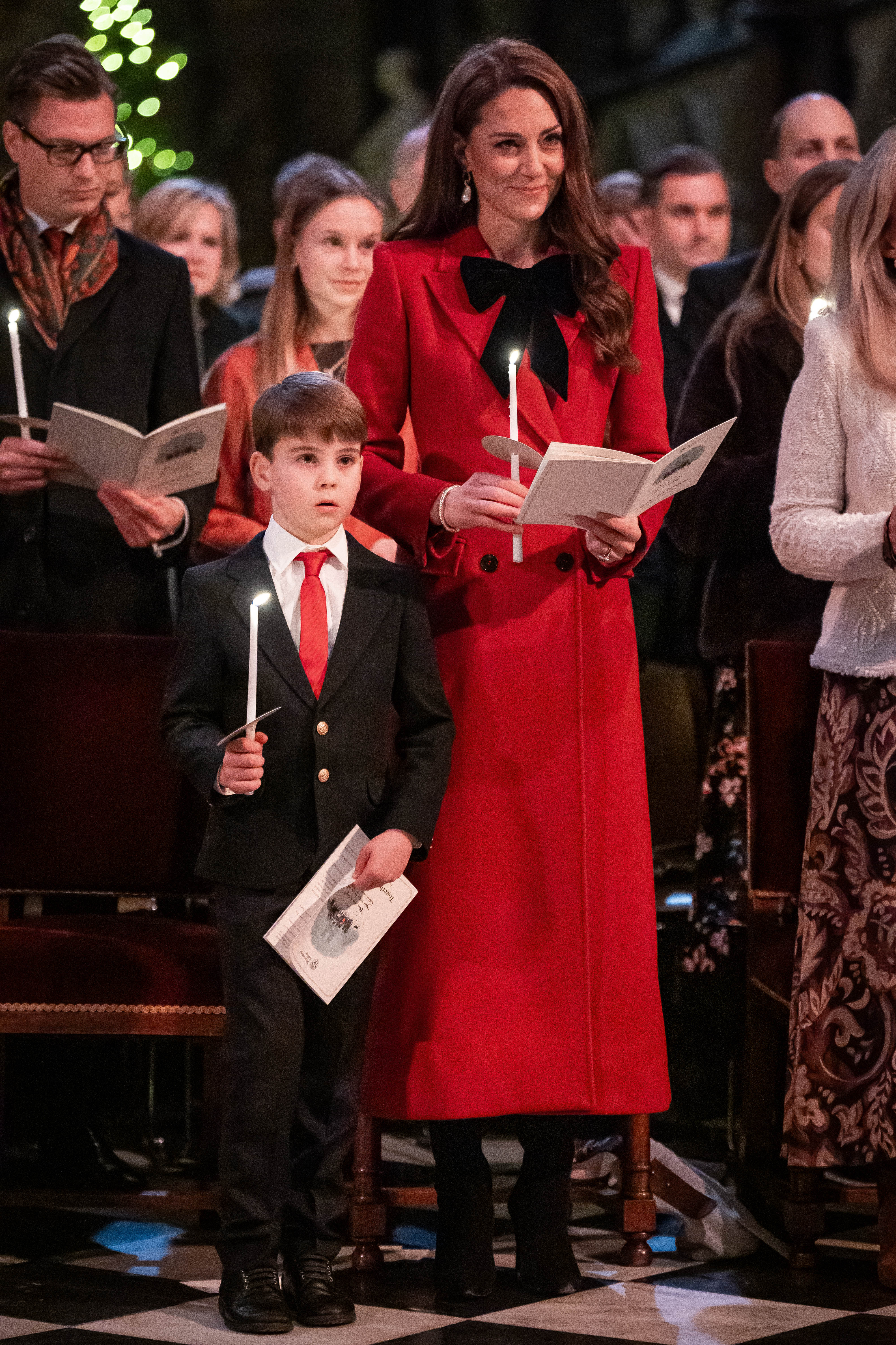 Kate Middleton wearing a red coat holding a candle standing next to Prince Louis in church