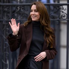 Kate Middleton wearing a black turtleneck and brown blazer smiling and waving in front of a metal fence