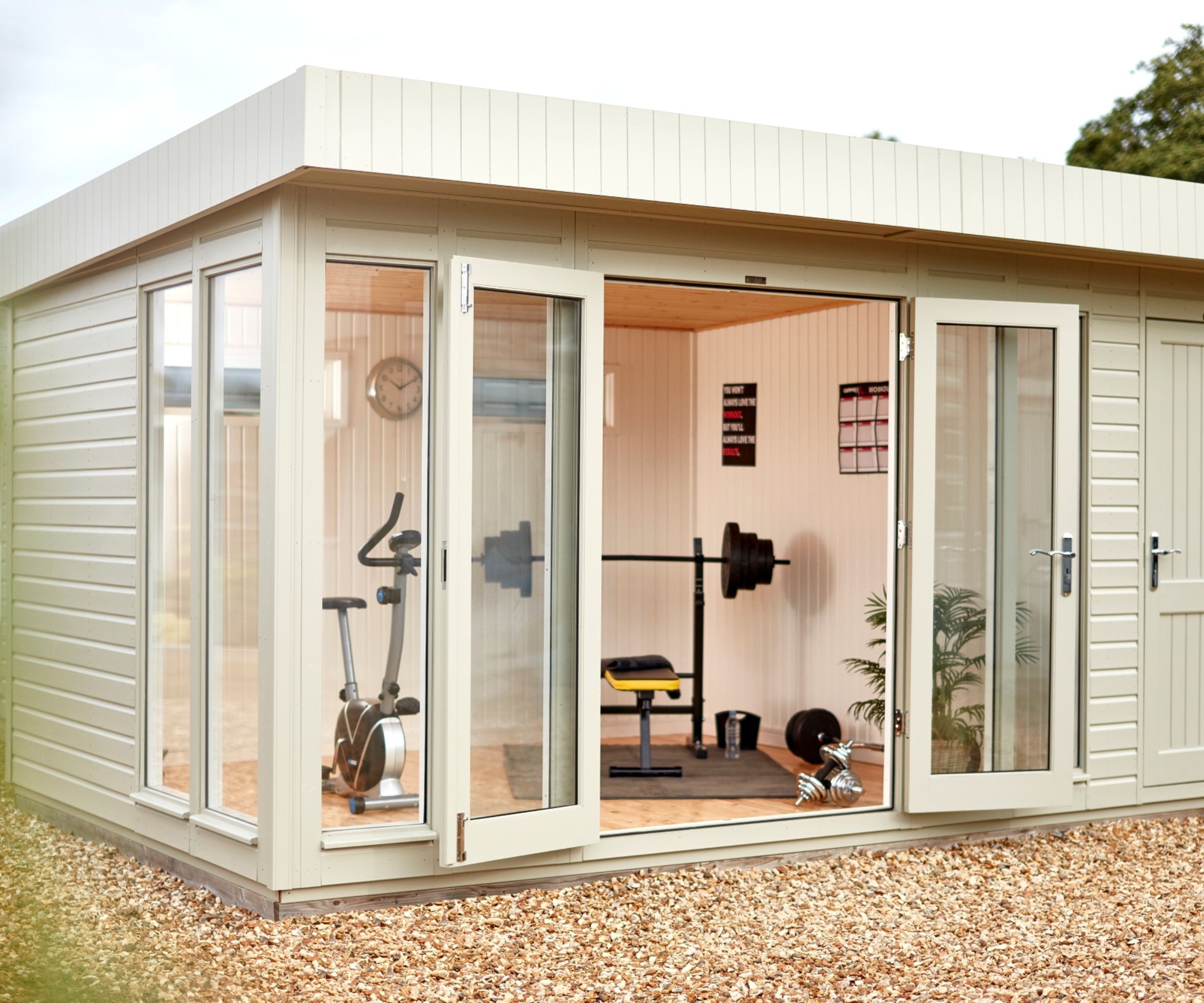 Cream garden building with open glass doors showing exercise bike, weight bench, barbell, and dumbbells inside.