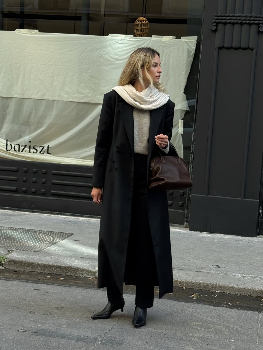 A woman wears a black coat and pants with a beige sweater, cream scarf, brown handbag and leather boots.