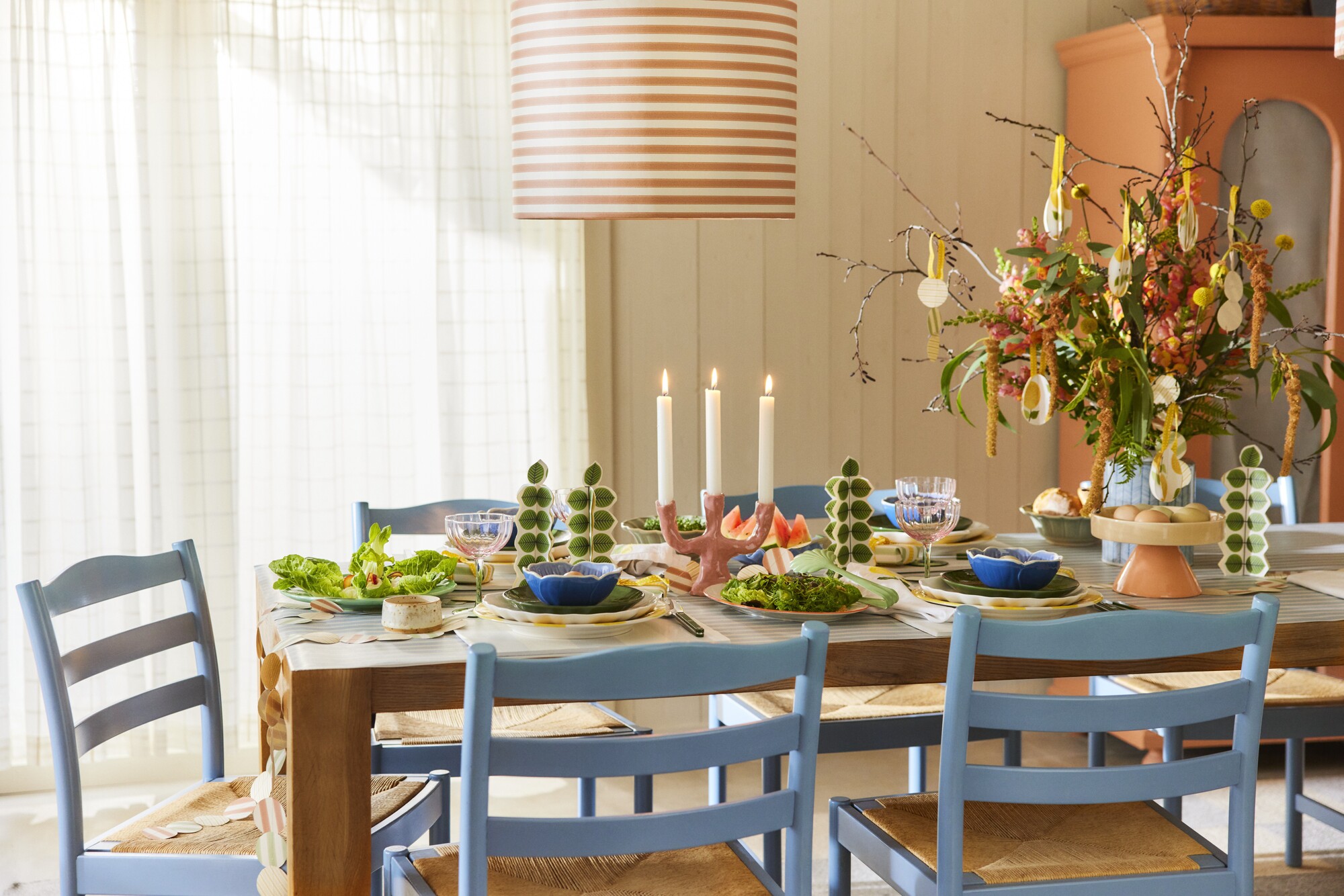 Image of a bright, airy dining room with a wooden table that has eight light blue and wicker chairs around it. The table is set for a spring dinner party.