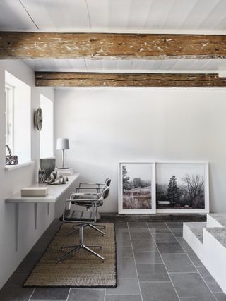 Study in the Vipp Farmhouse with two aluminium office chairs by the window, black and white framed photographs on the floor and visible wooden beams on the ceiling