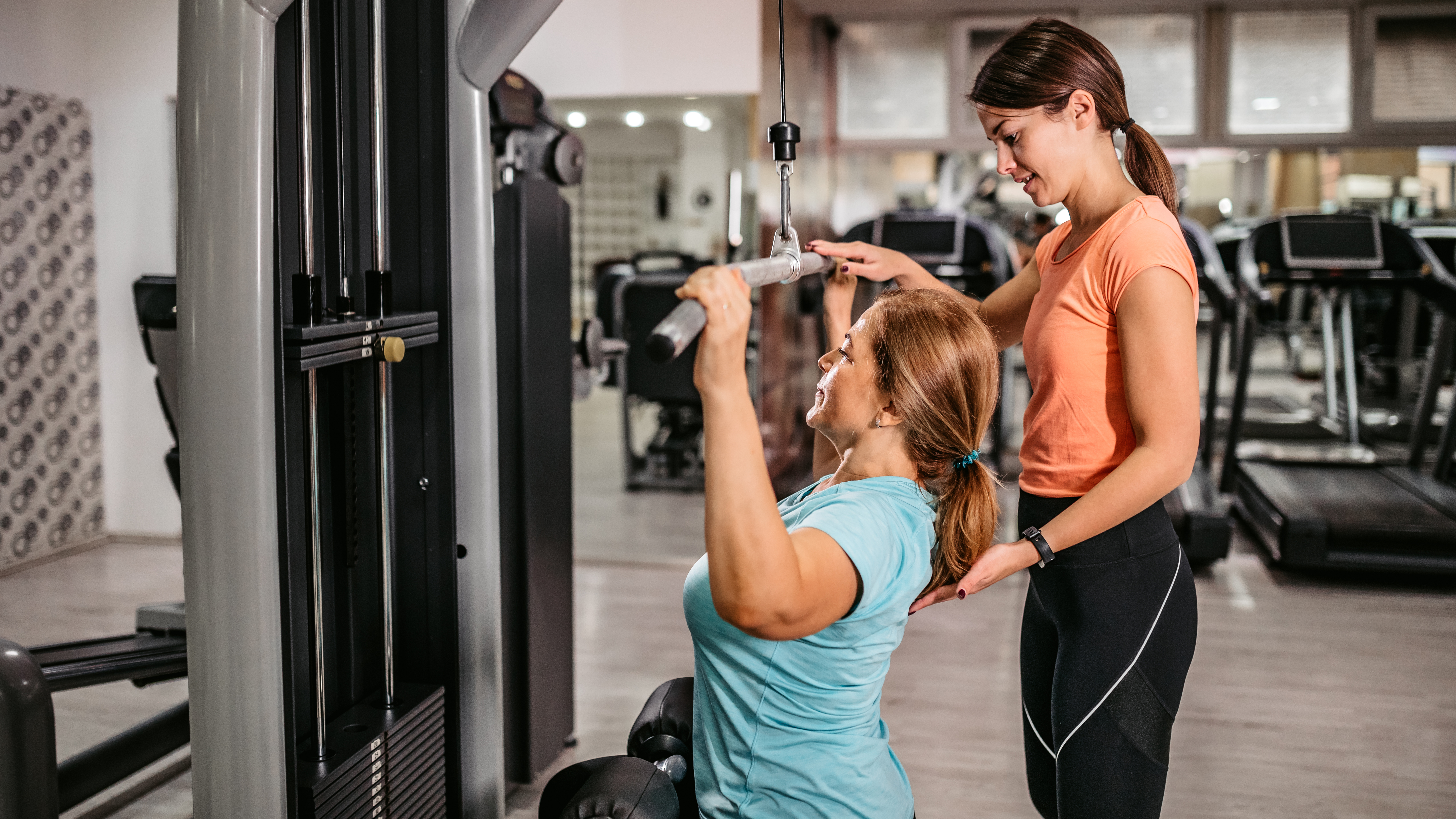 A trainer helps a woman at a gym practice a lateral pull-down on a machine. The trainer is gently touching the woman&#039;s back as she pulls the bar down to her head.