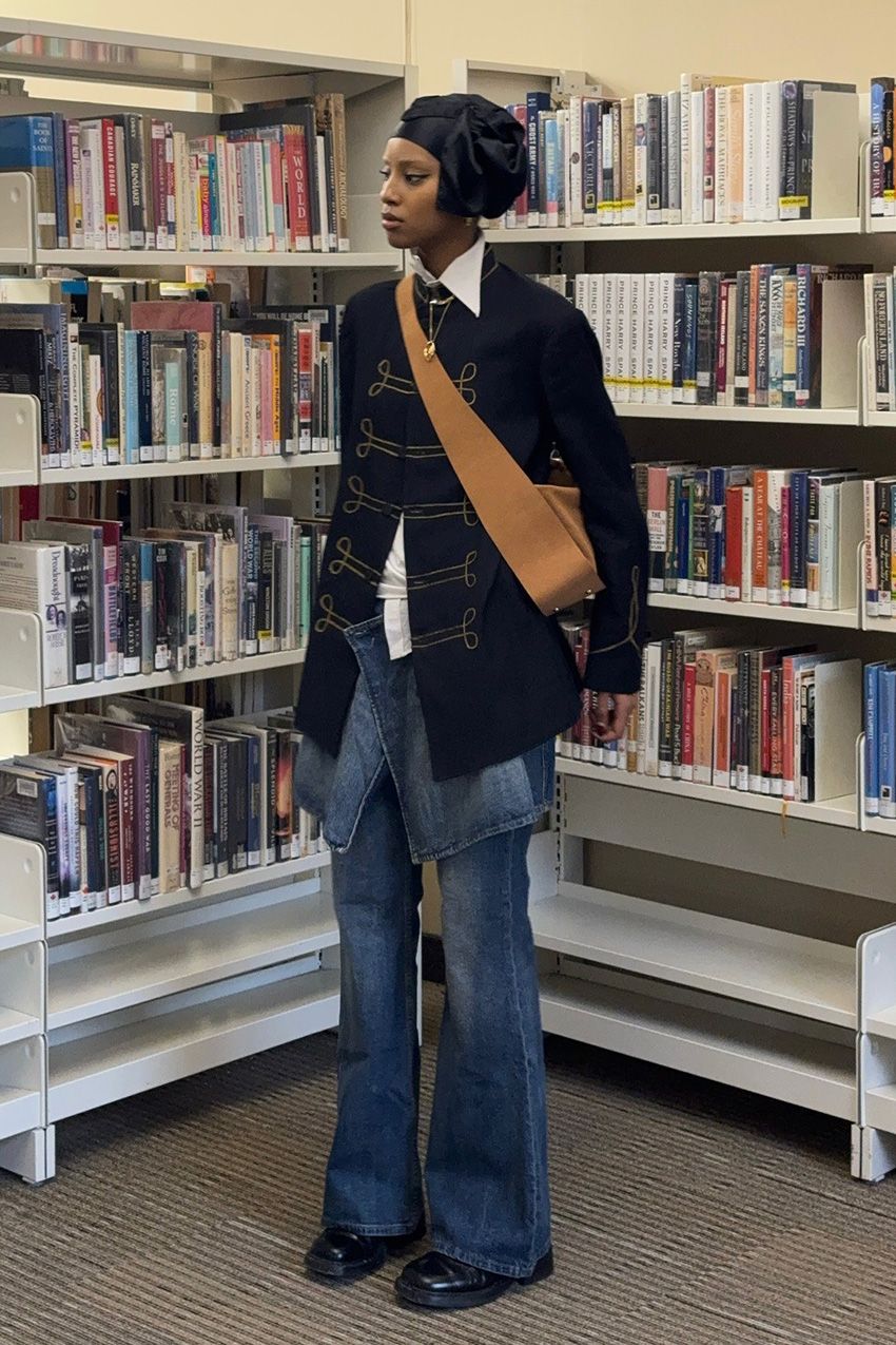 A woman standing inside a library wearing a navy military jacket over a white button-down shirt, a denim skirt over wide-leg jeans, black ankle boots, and a light brown crossbody bag.
