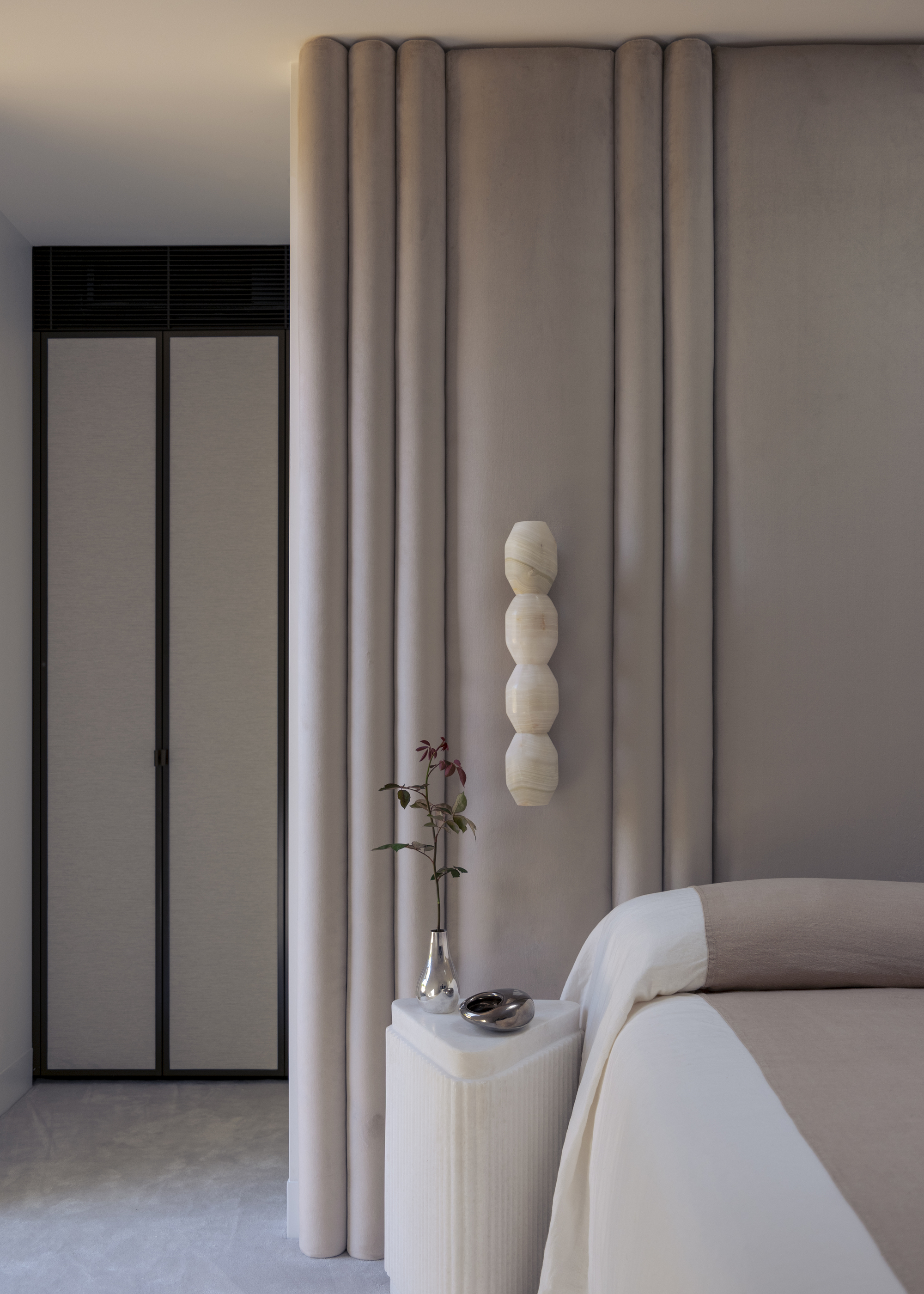 Image of a dusty pink bedroom with fabric on the walls and soft off-white carpeting. The bed has a minimal bedspread and there is a white sconce light and a side table.