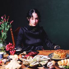 michelle zauner of japanese breakfast wears a black silk dress and sits at a table with lots of food 