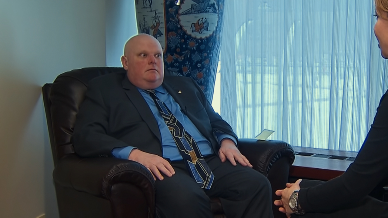 Rob Ford sitting in a chair in a suit, with a bald head, looking sick in Trainwreck: Mayor of Mayhem