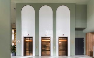Three gold hotel elevator doors, with white arched tiles around them on a grey high wall, a potted plant and a black door on the right of them.
