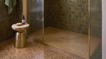 Warm beige shower tray with olive-green wall tiles, brass-framed glass screens, and a pink terrazzo floor beneath a plaster arch.