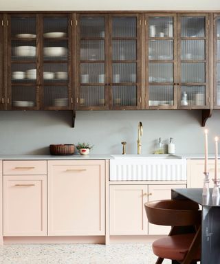 A kitchen with light pink bottom cabinets and dark wood top cabinets, a farmhouse sink with gold tap