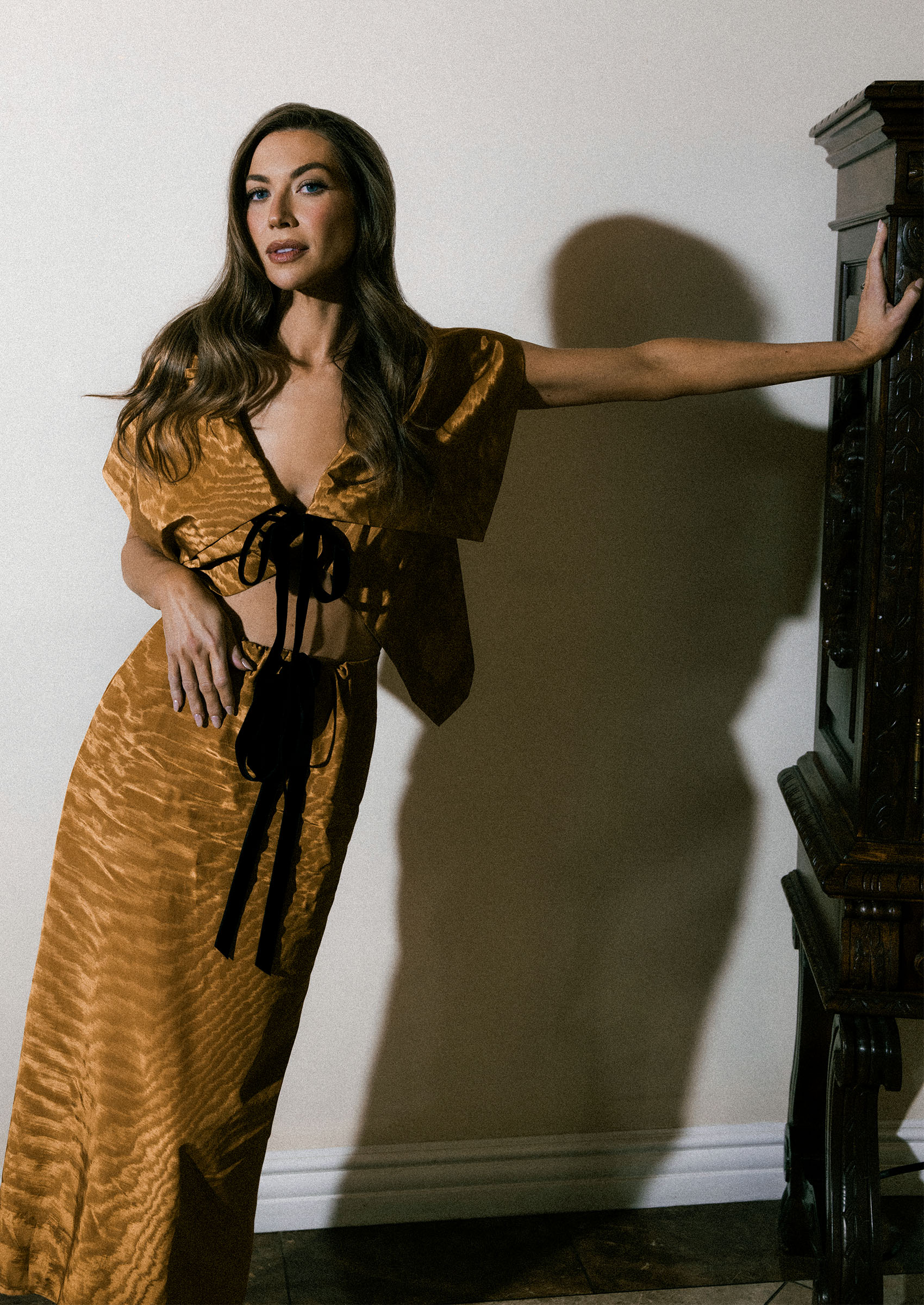 stassi schroeder poses wearing a brown set with black ties as she leans against a wall in an editorially shoot