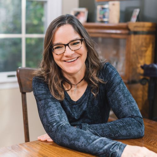 Jen is a white woman with medium length light brown hair. She's pictured wearing a long sleeve blue snake-skin pattern top, and black glasses, with a big smile for the camera