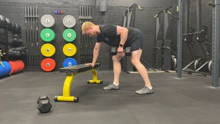 Fitness writer Harry Bullmore performs a single-arm bent-over dumbbell row in a gym. His right arm rests on a weight bench as he maintains a staggered stance and hinges forwards at the hips, so that his torso is parallel to the floor. In his right hand he holds a dumbbell. His right elbow is bent and close to his side. Behind him we see weight plates and gym equipment.