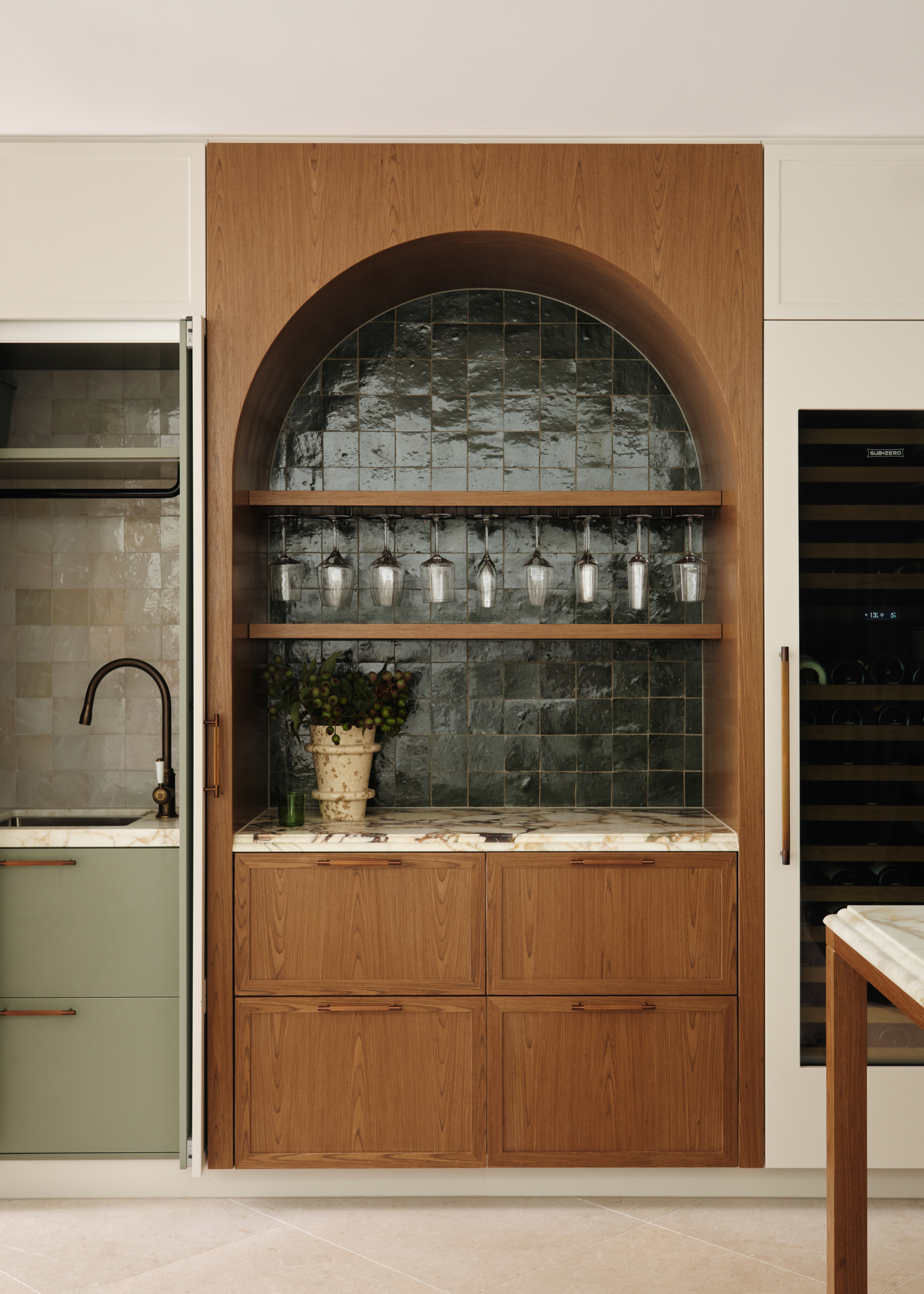 A kitchen with an arched in-set bar area with floating shelves featuring glassware, a vase of flowers on the marble countertop and wood drawers by a wine fridge