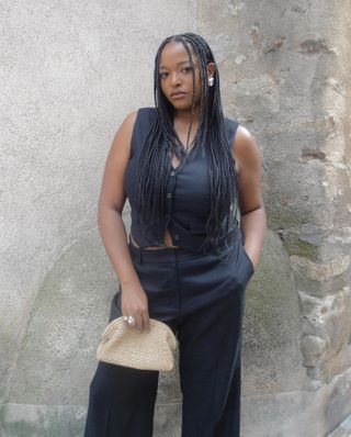 woman wearing black V-neck vest, black trouser pants, raffia clutch bag, silver earring, and ring and standing against stone wall