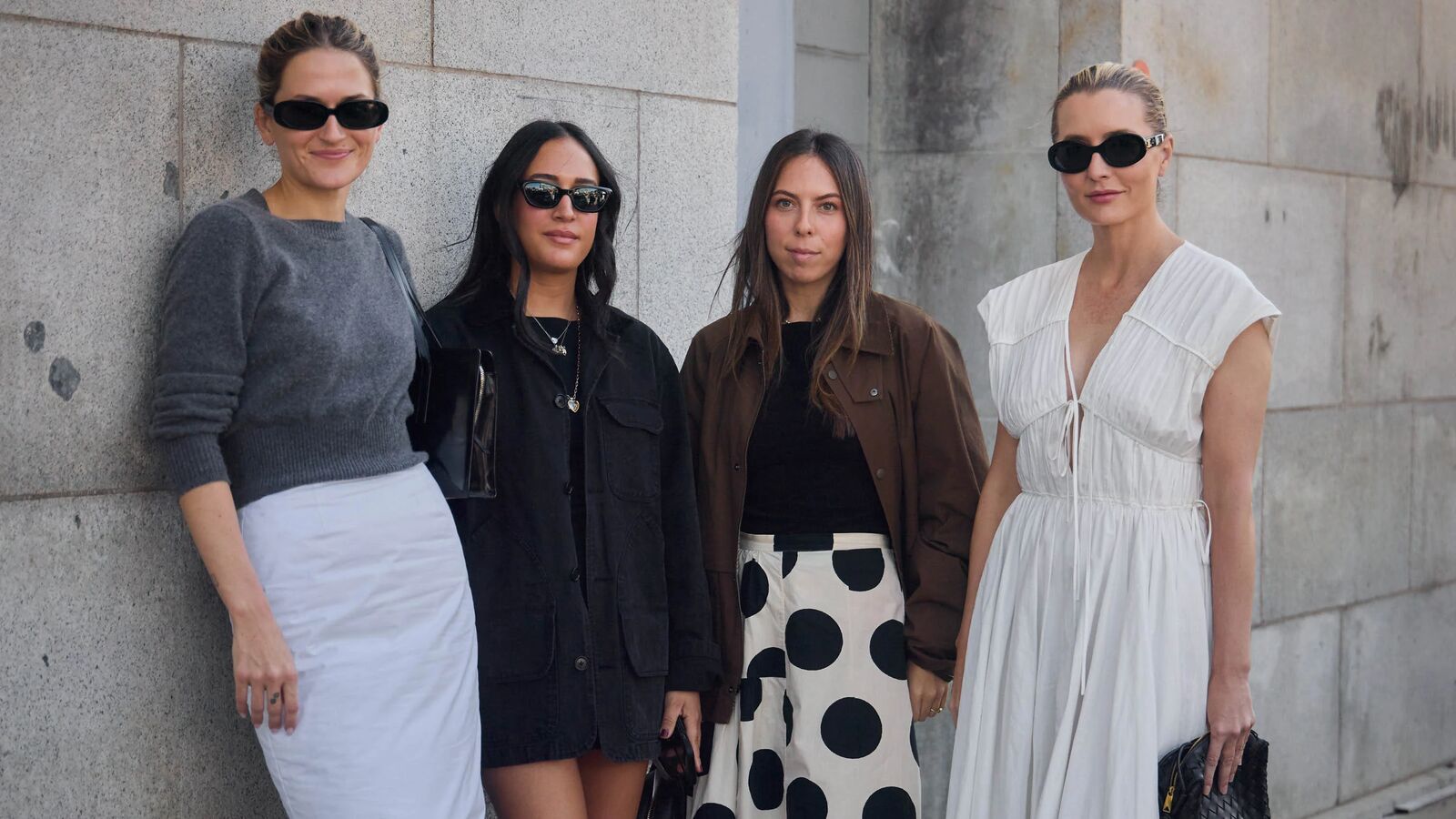 Four women pose for a picture. Eliza wears gray sweater and white pencil skirt. Nicole wears a black dress. Lauren wears a polka dot skirt, black top, and brown bomber. Kristen wears white dress.