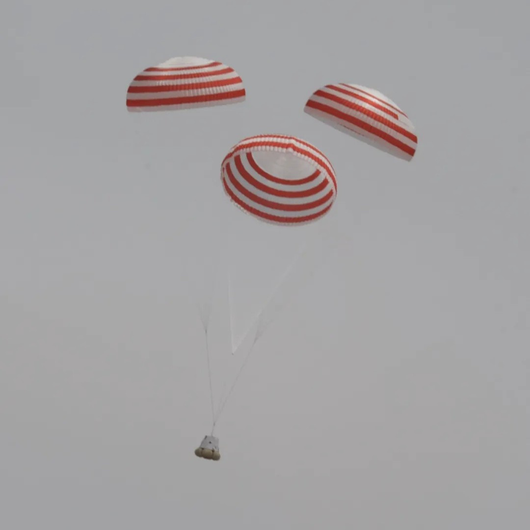 a space capsule falls against a grey sky, lowered by three white and red striped parachutes.
