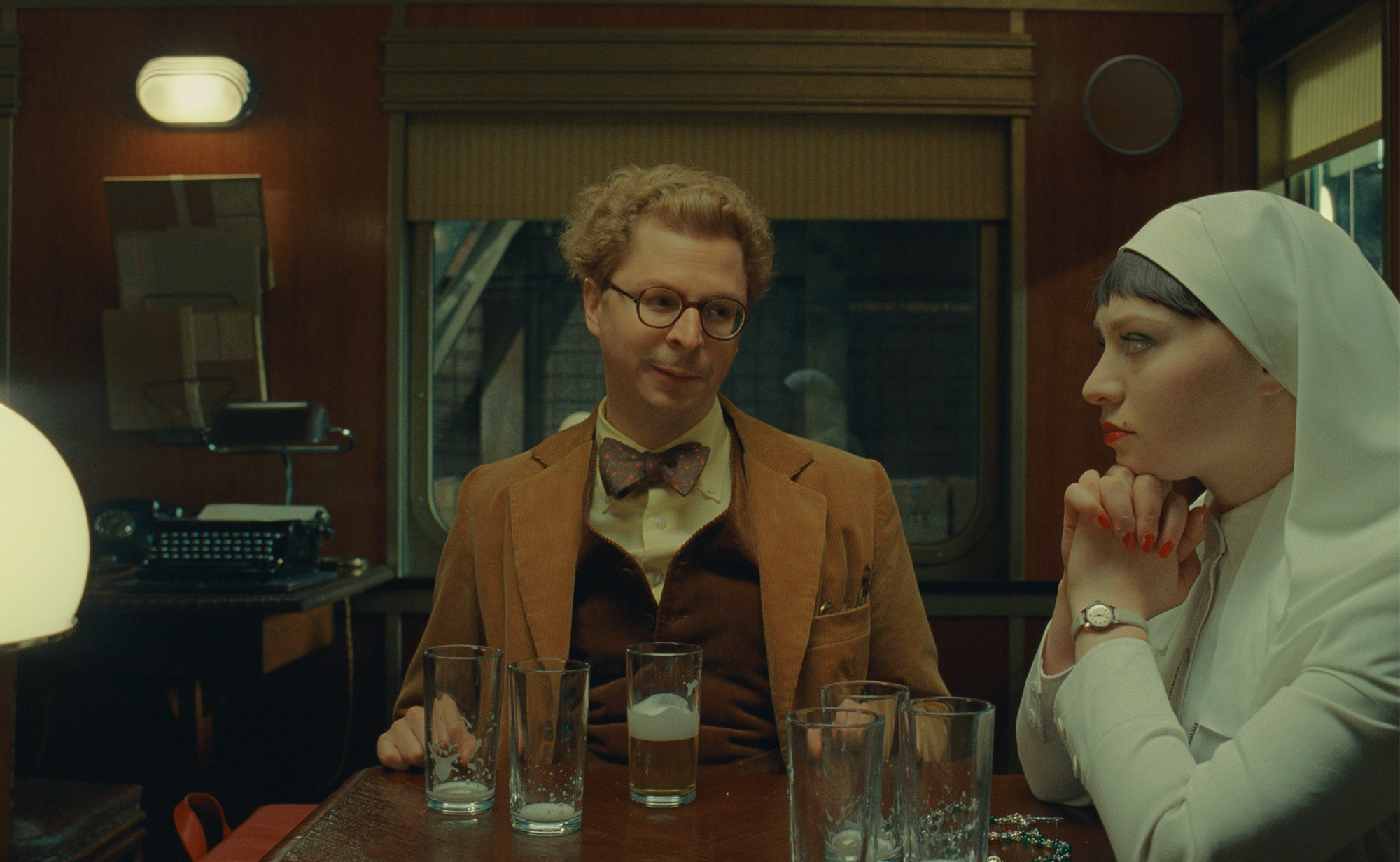 Still from the film The Phoenician Scheme showing Michael Cera and Mia Threapleton sitting next to one another at a table full of beer glasses.